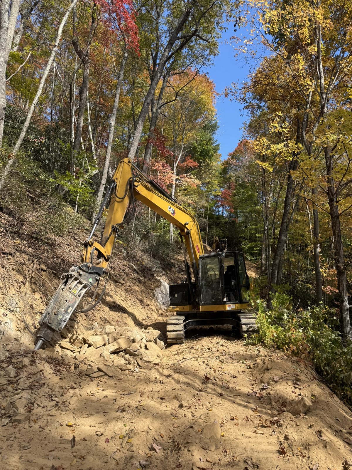 Excavator with a hydraulic rock hammer breaking stone on a slope in Western NC